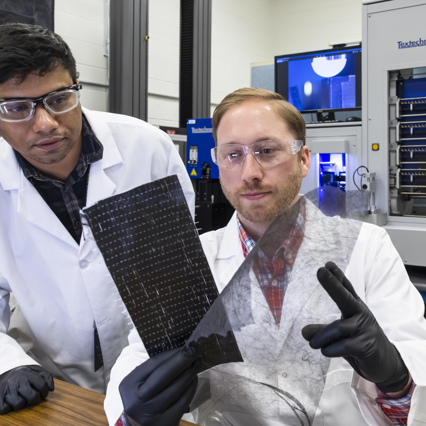 Two ORNL researchers inspect carbon fiber materials - one black rectangular sheet and one see-through sheet of film. 